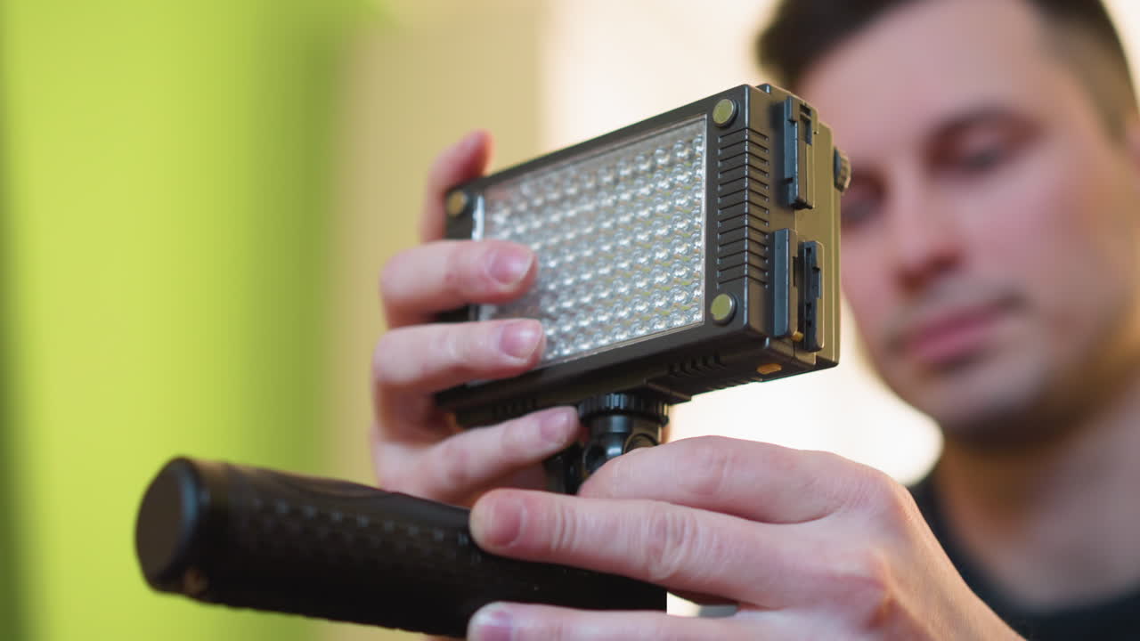 Studio guy adjusting camera light panel with focus on hands. Camera and microphone setup visible in background. Preparing equipment for filming in a bright, modern studio environment with green accents