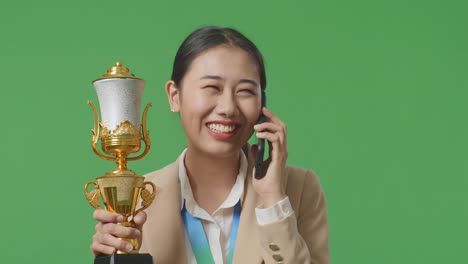 close up of asian business woman in a suit with a gold medal holding a gold trophy and talking on smartphone on green screen background in the studio