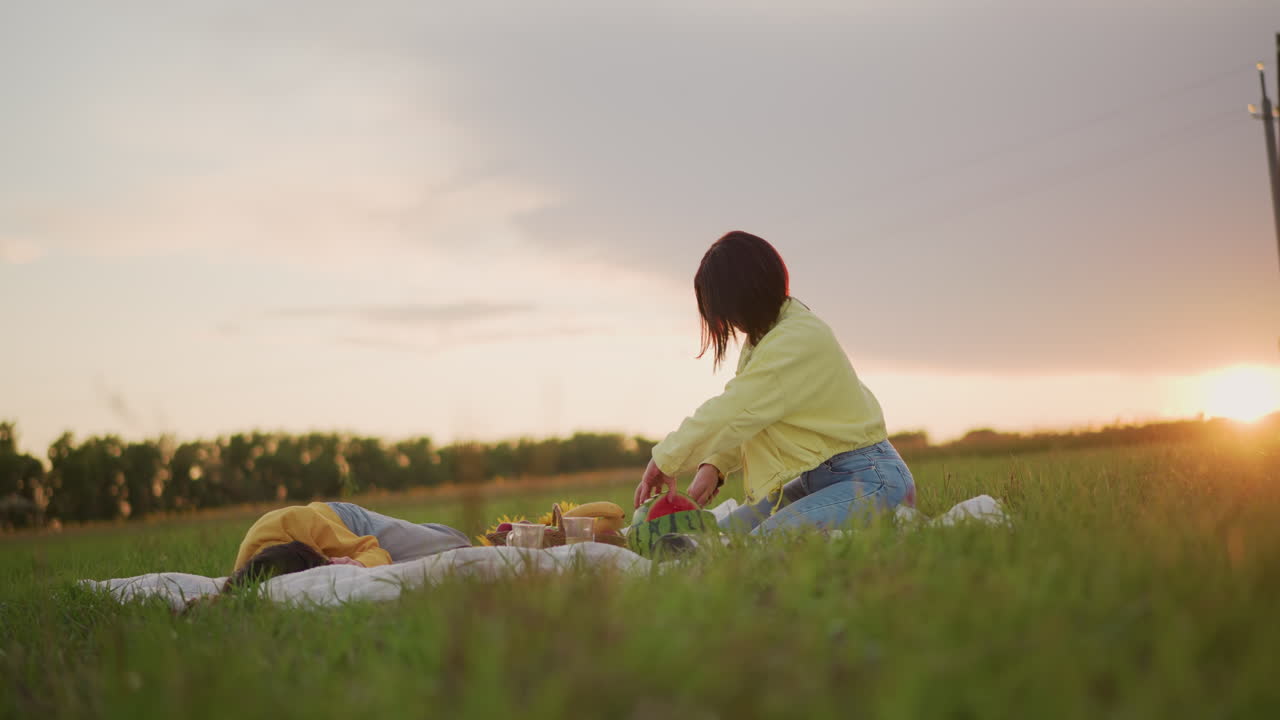 해질녘 피크닉에서 흑인 여성이 누워 있는 친구 옆 담요 위에서 수박 조각을 건네고 있고, 태양 플레어와 따뜻한 햇살, 즐거운 미소와 캐주얼한 데님, 피크닉 바구니와 안경, 편안한 초원 분위기가 연출되고 있어요.
