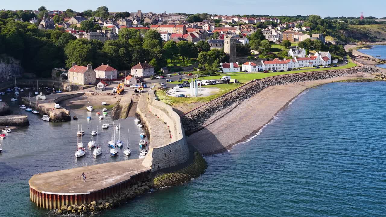 Drone sobrevoa vila litorânea, porto de pedra, barcos atracados e casas à beira-mar em plena luz do dia