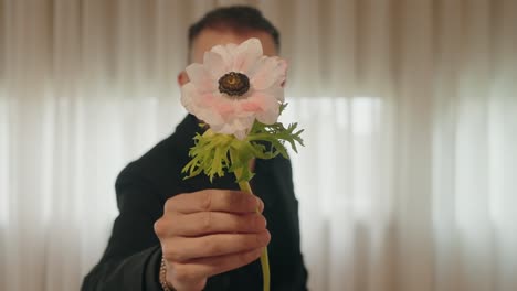 man in black suit offers flower with soft smile, focus on the flower