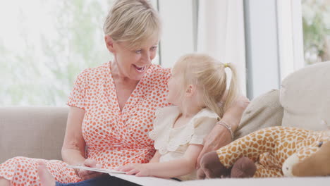 grandmother sitting on sofa with granddaughter at home reading book together