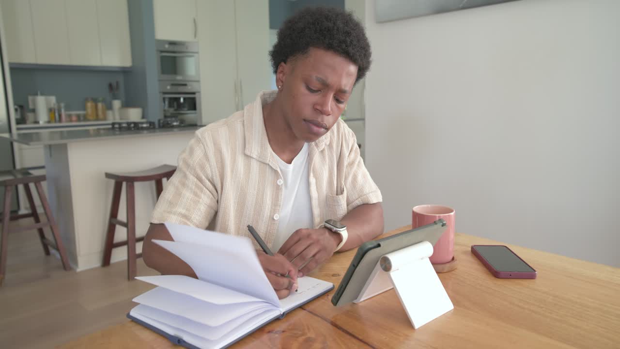 Uomo afroamericano sui vent'anni che studia a un tavolo di legno, consultando un tablet e scrivendo su una pila di quaderni.