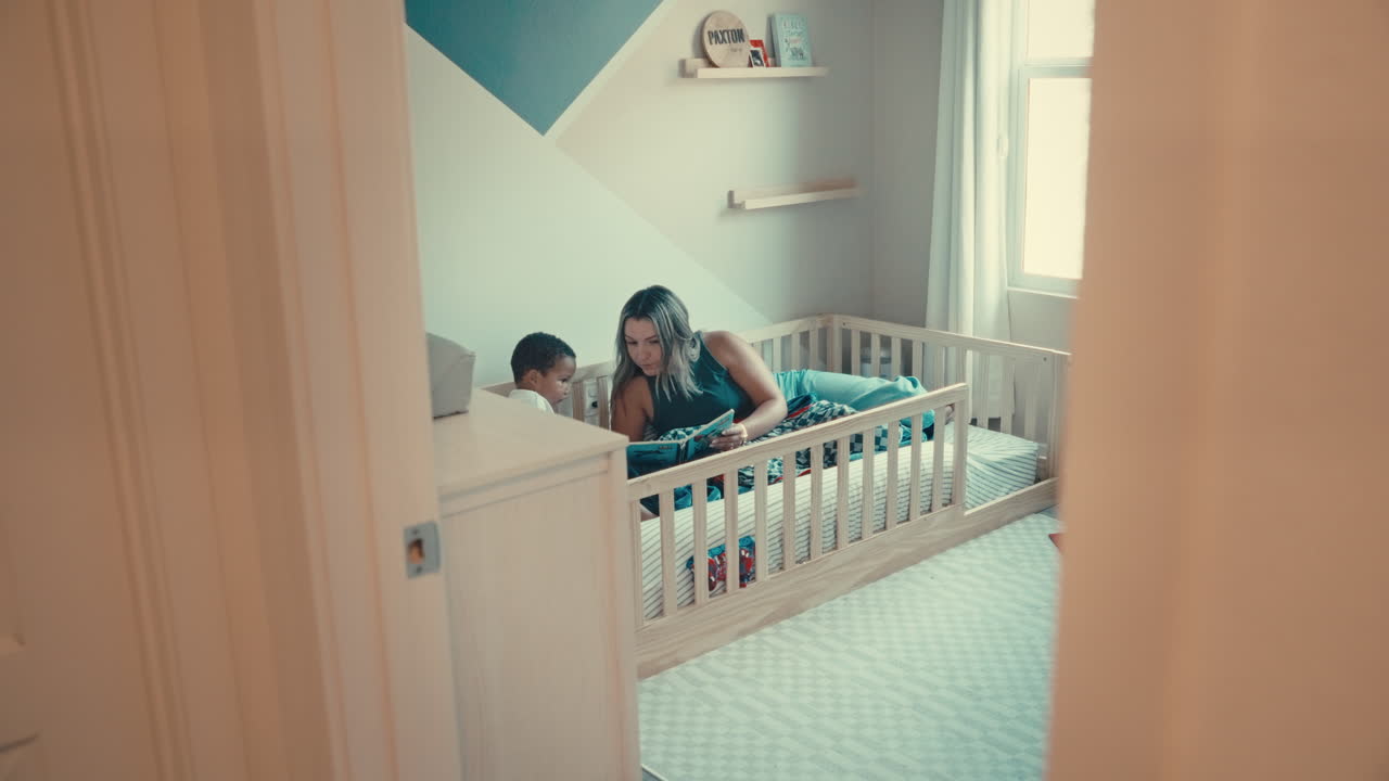 Wide shot from hallway shows Hispanic mom reading to Black toddler in bed. Captures cozy nursery, parent child bonding, and warm, authentic home environment