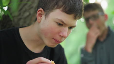 close-up of young boy wearing a black top enjoying a snack under a tree, he is focused on his food, while in the background, a blurred view of another person is also seen eating under a leafy tree