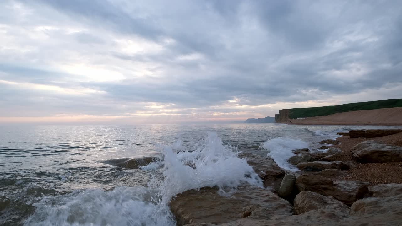 Gentle waves crashing on beach rocks on the Jurassic Coast, Dorset. Slow motion captures the ripples and splashes of the water bouncing off the rocks. Captured on a cloudy day with cliffs behind.