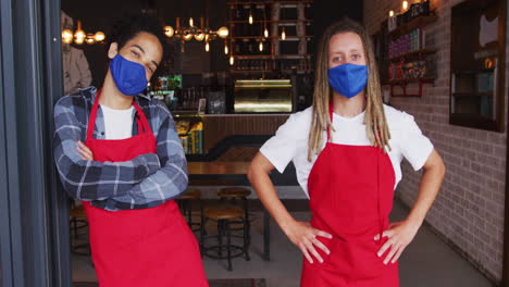 two diverse male baristas wearing face masks and aprons standing in doorway of cafe