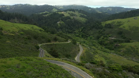 Airel-pull-back-shot-coming-over-the-crest-of-a-mountain-ridge-on-a-very-windy-road-with-cars-passing-by-in-northern-California