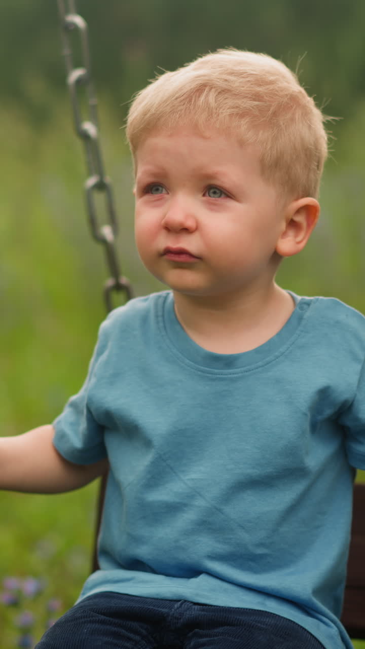 Upset weepy little kid boy sits on wooden swings on playground against high lush grass at countryside on spring day closeup slow motion