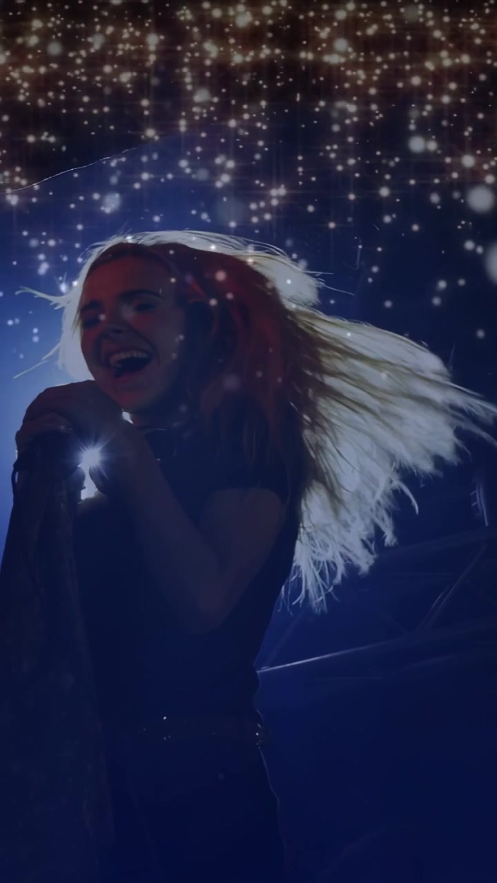 Video verticale: Cantante che si scuote i capelli, si avvicina al microfono su un palco musicale, cantando tra stelle di luce blu.