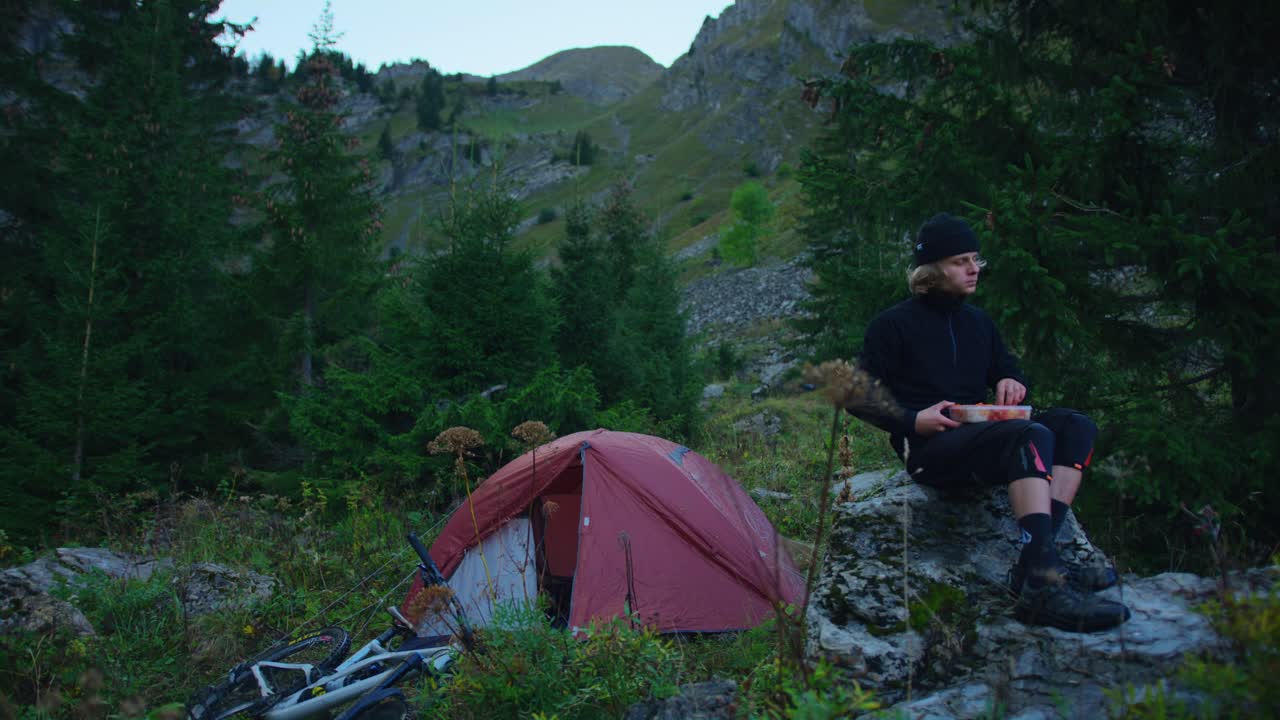 un ciclista de montaña está comiendo junto a su tienda en un valle alpino