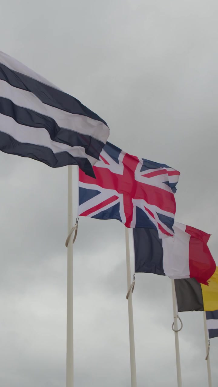 Multiple national flags wave vigorously on tall poles under cloudy skies at a coastal memorial