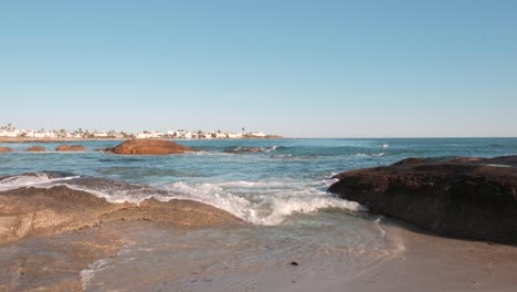 Vista-De-Las-Olas-Del-Océano-Rompiendo-En-Las-Rocas-En-Primer-Plano-Con-Casas-En-La-Distancia