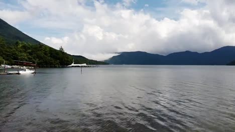 Vuelo-Bajo-Sobre-Un-Hermoso-Lago-En-Un-Día-Ventoso-Con-Nubes-Esponjosas-Y-Montañas-En-El-Fondo