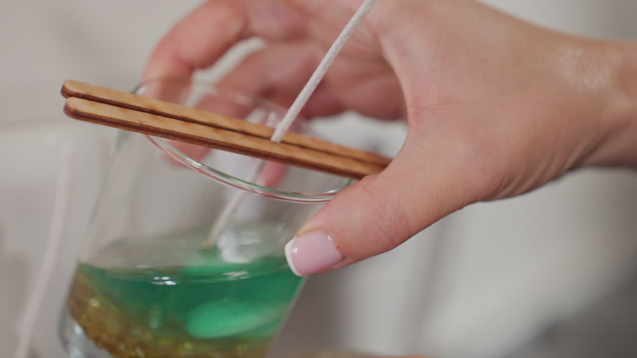 Close-up of glass cup filled with green liquid, golden bead, and seashell, wick placed carefully in cup, with the cup resting on a burlap sack beside two other cups in candle making process