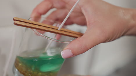 close-up of glass cup filled with green liquid, golden bead, and seashell, wick placed carefully in cup, with the cup resting on a burlap sack beside two other cups in candle making process