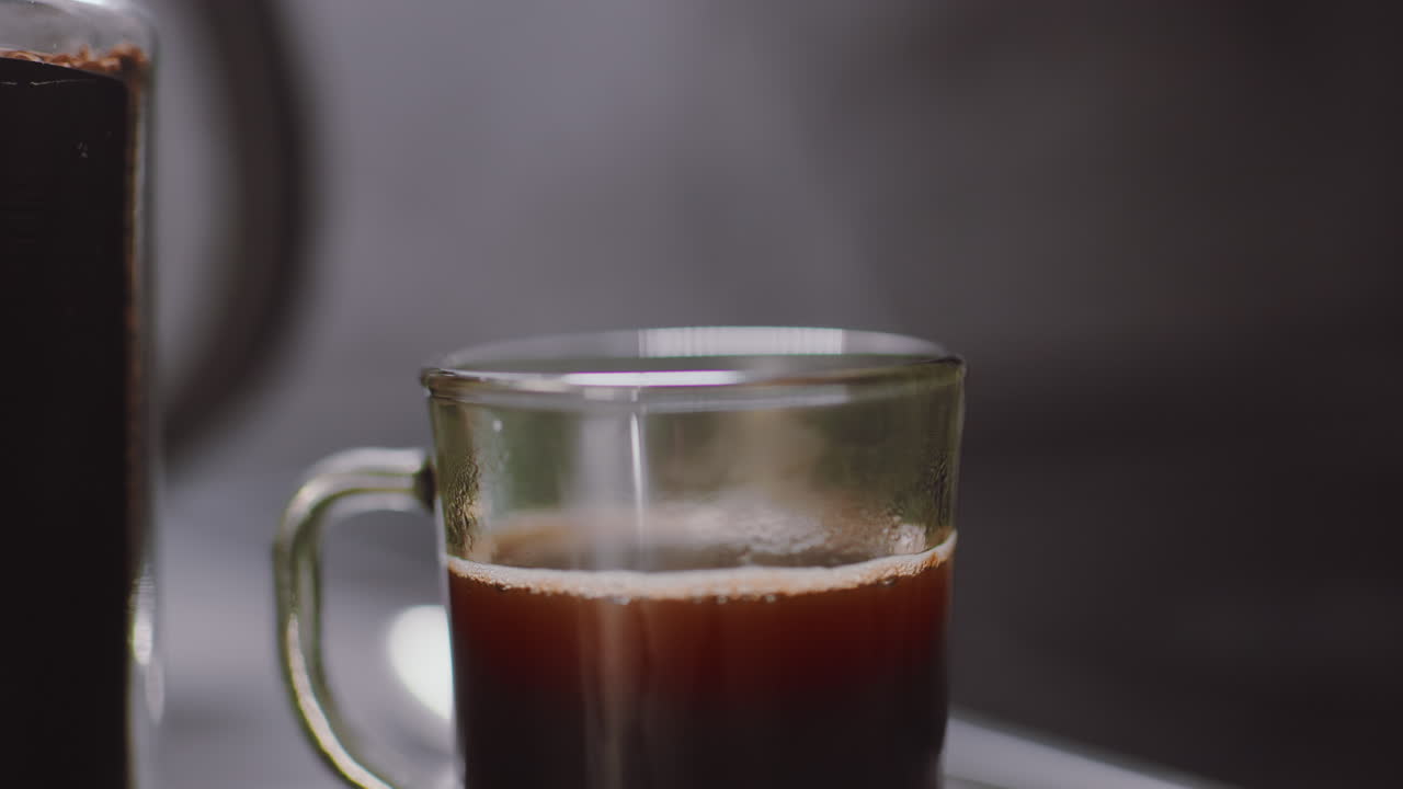 Close up of glass mug filled with hot dark coffee showing gentle rising steam, with soft morning lighting and part of coffee jar visible on counter