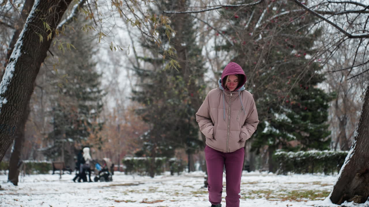 Une jeune fille saute joyeusement dans un parc enneigé en hiver, vêtue d'un sweat à capuche chaud et d'un manteau, des arbres enneigés l'entourent tandis que des personnes en arrière-plan poussent un trotteur pour bébé.