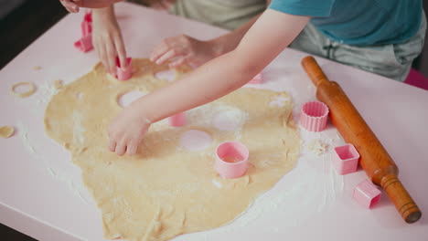 Children arranging cutout dough shapes while focusing on cutting new designs using pink plastic cutters on floured table with wooden rolling pin nearby during fun home baking session