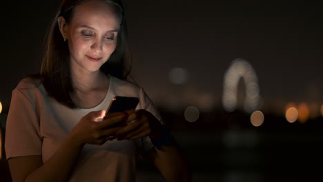 a young woman looks into the smartphone and writes text messages on the internet against the backdrop of the night city. girl businessman working on vacation remote work via mobile phone. gadget addiction