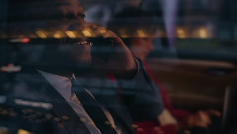 handsome businessman and beautiful businesswoman riding on the backseat of a car in the evening. man makes a phone call, woman works on a laptop. camera shot made from outside the car.