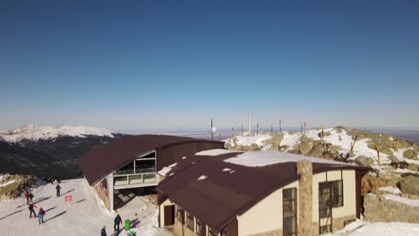 Raising-up-shot-from-behind-of-a-ski-station-as-skiing-people-passing-by-revealing-an-endless-winter-scenery-with-a-mountain-range