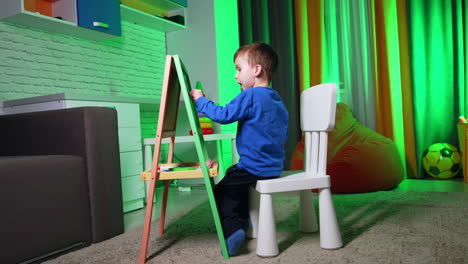 Little toddler kid is focused on the drawing. Baby boy uses chalk to draw on the blackboard.
