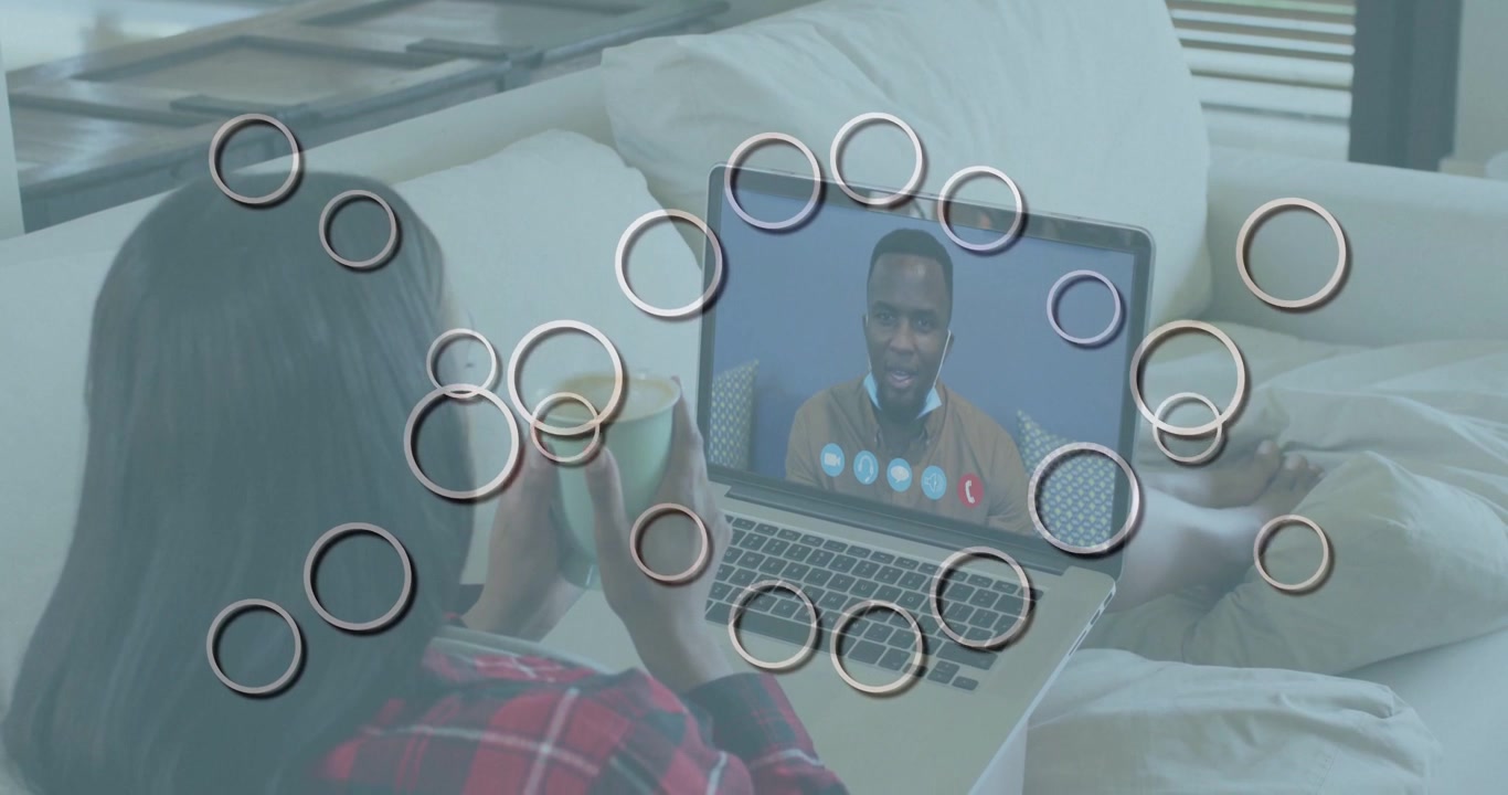 Donna seduta a letto con tazza e laptop, in videochiamata con un uomo che parla, icone e cerchi animati in chat.