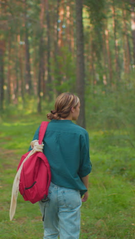 vista trasera de una mujer caminando por un tranquilo bosque iluminado por el sol, mochila roja colgada sobre el hombro, camisa verde mezclada con la naturaleza, la luz del sol se filtra a través de los árboles y la hierba alta