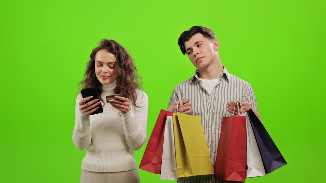 한 남자가 피곤하고 쇼핑백을 들고, 한 여자가 온라인 구매를 위해 행복하게 지불하고, 그들은 녹색 배경에 서 있습니다.