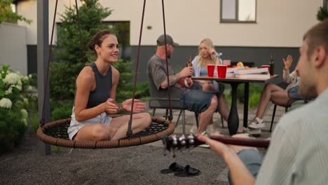 über-Die-Schulter-Klatscht-Ein-Glückliches-Brünettes-Mädchen-Und-Singt-Lieder-Zu-Der-Melodie,-Die-Ihr-Freund-Auf-Der-Gitarre-Spielt,-Während-Einer-Party-Mit-Seiner-Firma-Im-Hinterhof-Eines-Landhauses