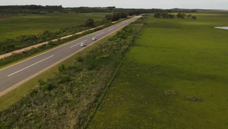 Two-white-vans-driving-on-asphalt-road-surrounded-by-rural-green-fields-in-Uruguay---aerial-tracking-shot