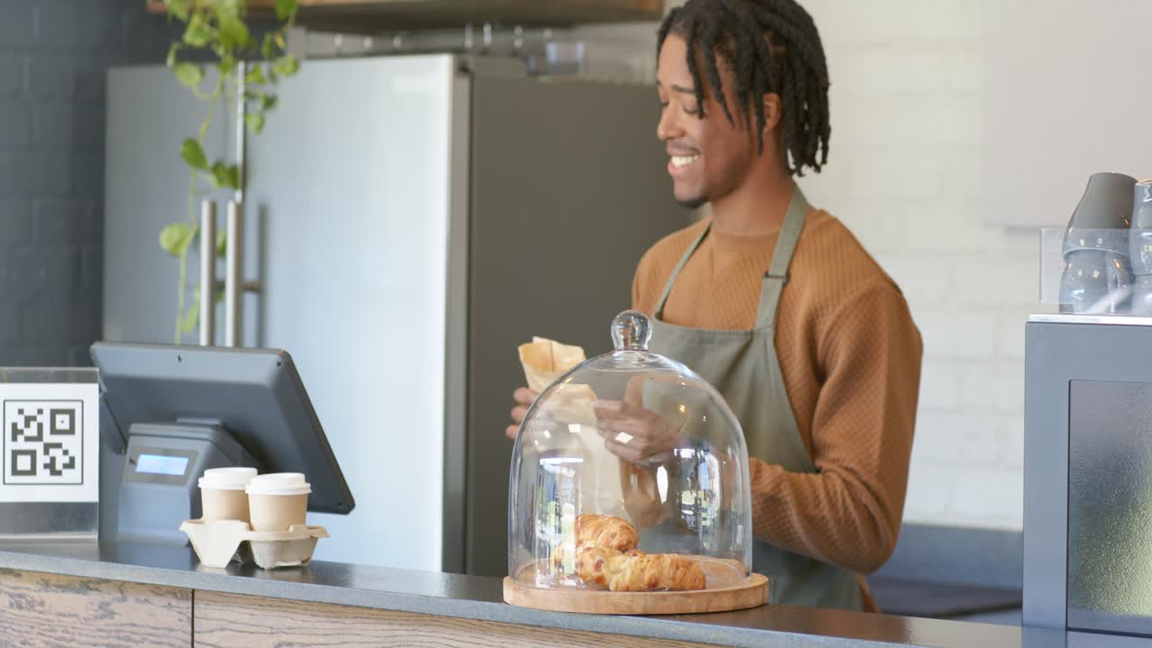 Ao ver o cliente, barista afro-americano de avental acenando e ensacando croissants em cafeteria com espaço livre