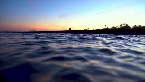 Wasser-Fließt-Mit-Voller-Geschwindigkeit,-Während-Man-Am-Horizont-Die-Schatten-Der-Menschen-Sieht,-Die-Sich-Am-Strand-In-Einem-Wunderschönen,-Orangefarbenen-Und-Farbenfrohen-Hintergrundlicht-Befinden