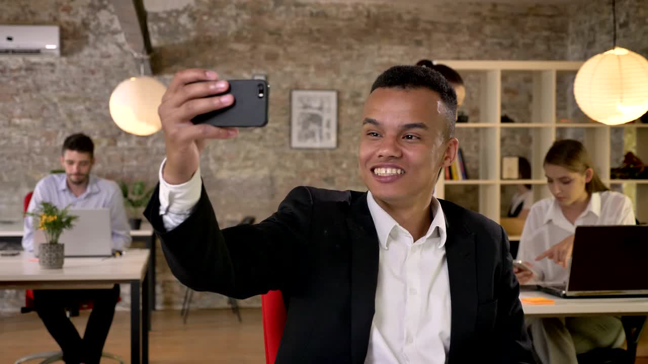 Young businessman is making selfie on smartphone in office, fools around, his colleagues are networking with technologies, working concept, business concept, communication concept