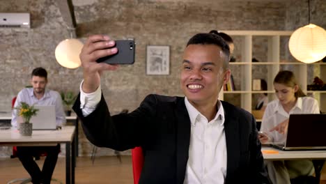 young businessman is making selfie on smartphone in office, fools around, his colleagues are networking with technologies, working concept, business concept, communication concept