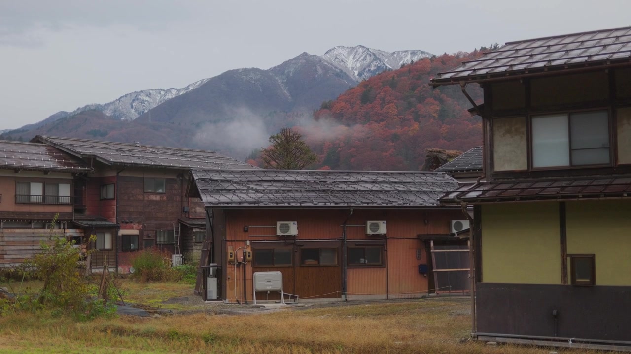 Klidná japonská vesnice s tradičními domy, podzimními kopci a zasněženými horami v pozadí. Malebná venkovská krajina s poklidnou venkovskou atmosférou.