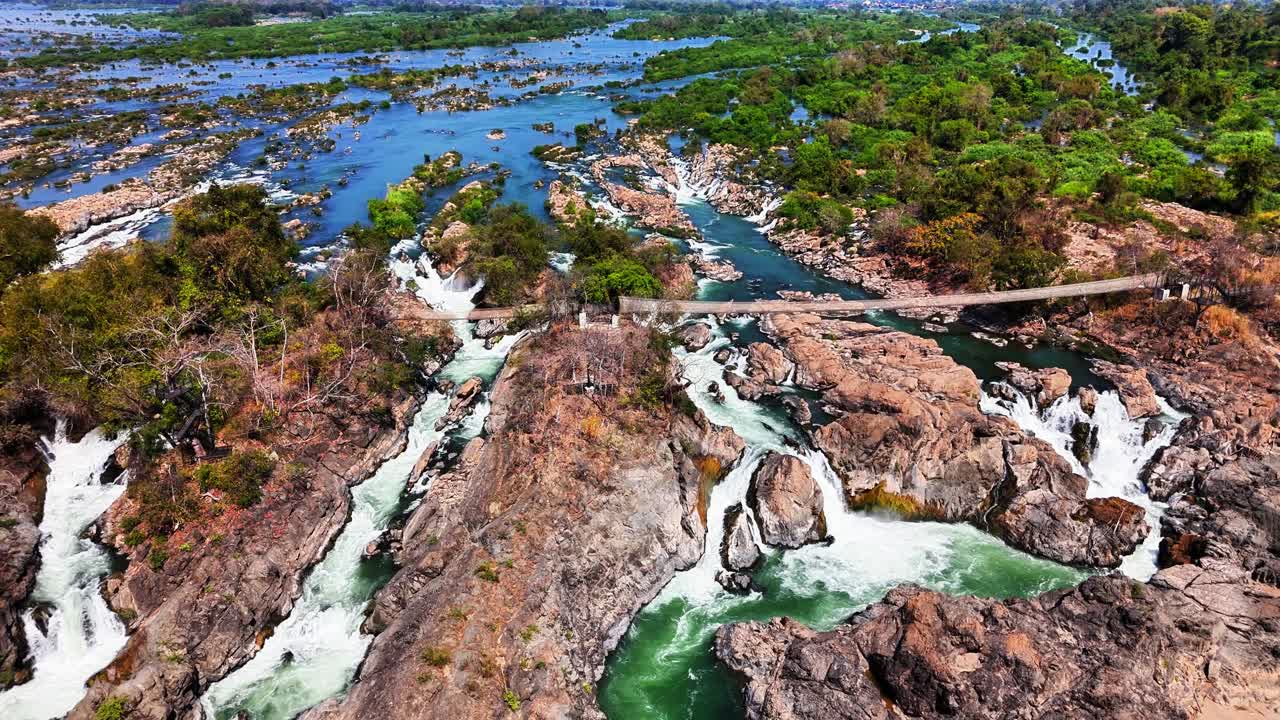 Drone view of the Mekong River splitting through rocky rapids and green islands at Don Det, Laos, with cascading waterfalls, fast-moving water, and a footbridge connecting lush riverbanks
