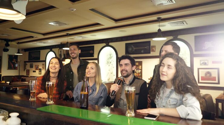 Giovani amici allegri cantano insieme in un karaoke bar mentre sono seduti al bancone e si divertono. Ridono, ballano e gesticolano godendosi la serata