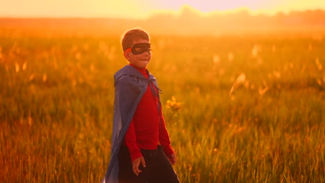 un niño disfrazado de superhéroe con una capa roja corre a través del césped verde contra el telón de fondo de una puesta de sol hacia la cámara.