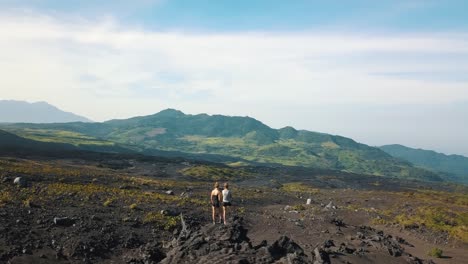 drone aerial rotating shot of two girls standing on a rock, looking to the distance near camping spot in pacaya volcano, guatemala