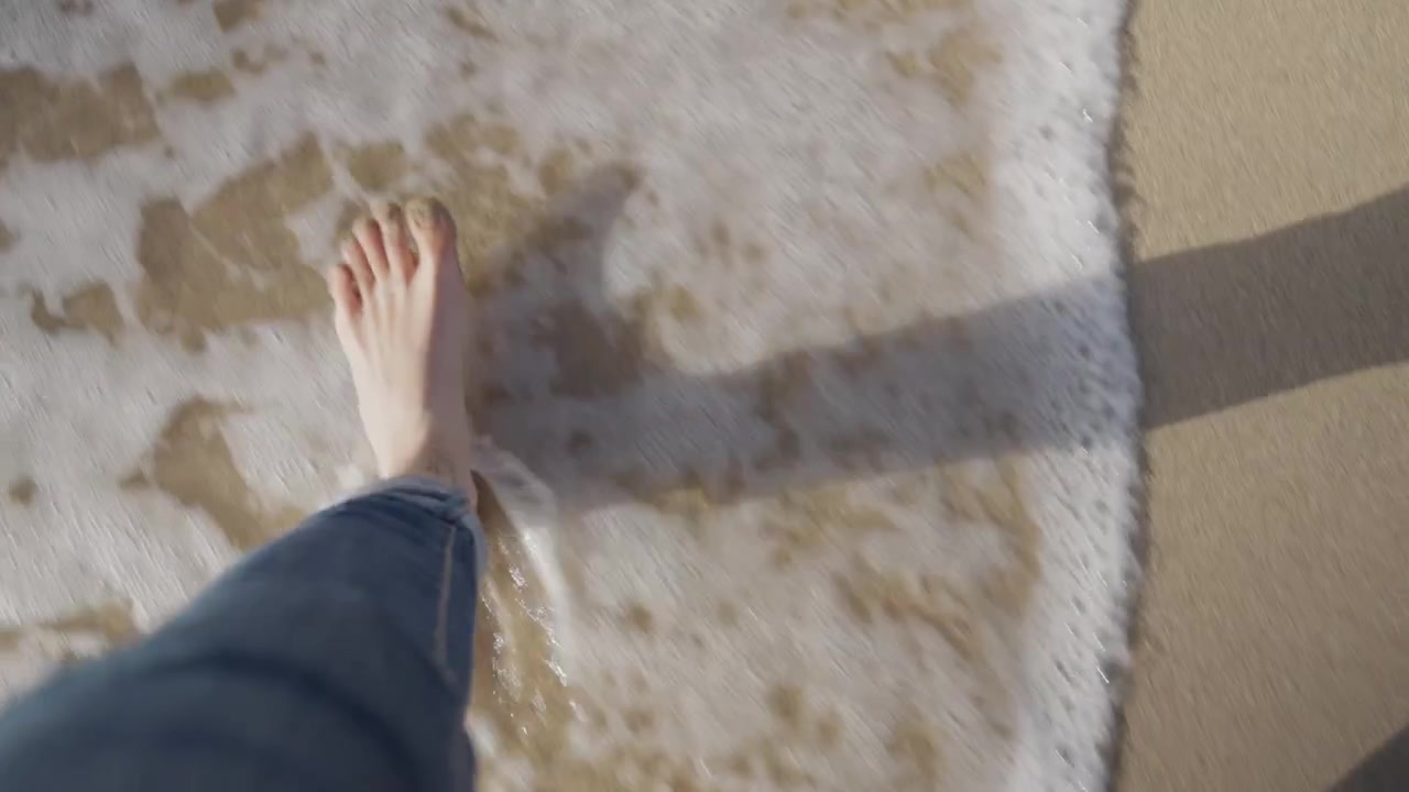POV top down shot of female feet walking on sandy beach as sea or ocean water washes her feet during tide. Peaceful bare feet stroll on shore during vacation holiday weekend. Relaxing paradise idyllic