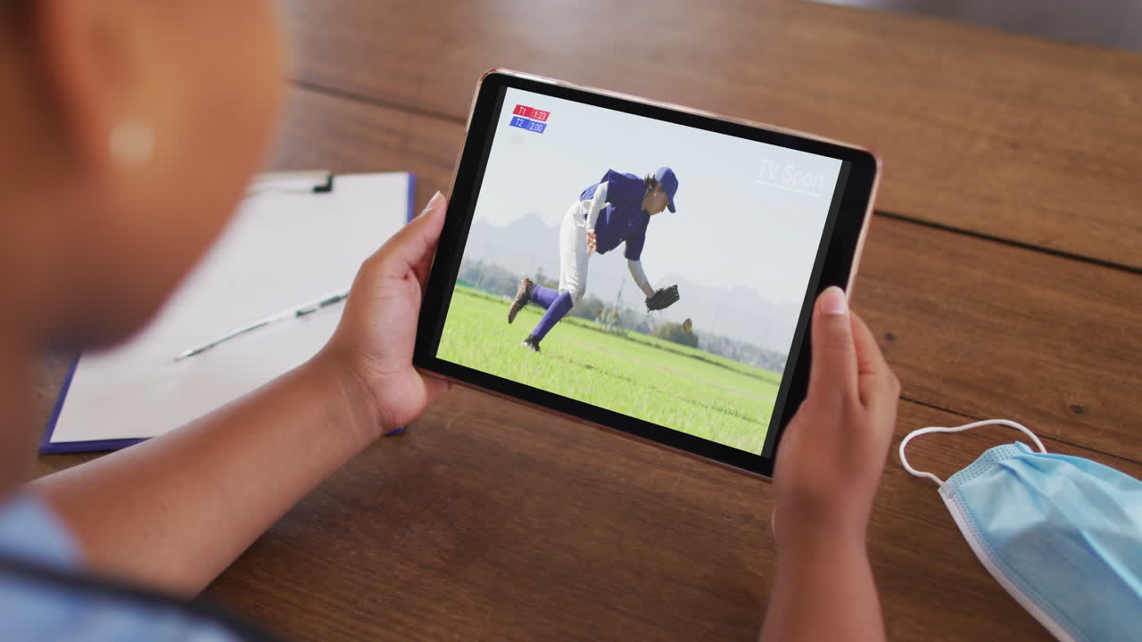 태블릿에서 야구 경기를 시청하는 얼굴 마스크와 함께 테이블에 앉아있는 여성의 복합