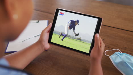 compuesto de una mujer sentada en una mesa con una máscara facial, viendo un juego de béisbol en la tableta