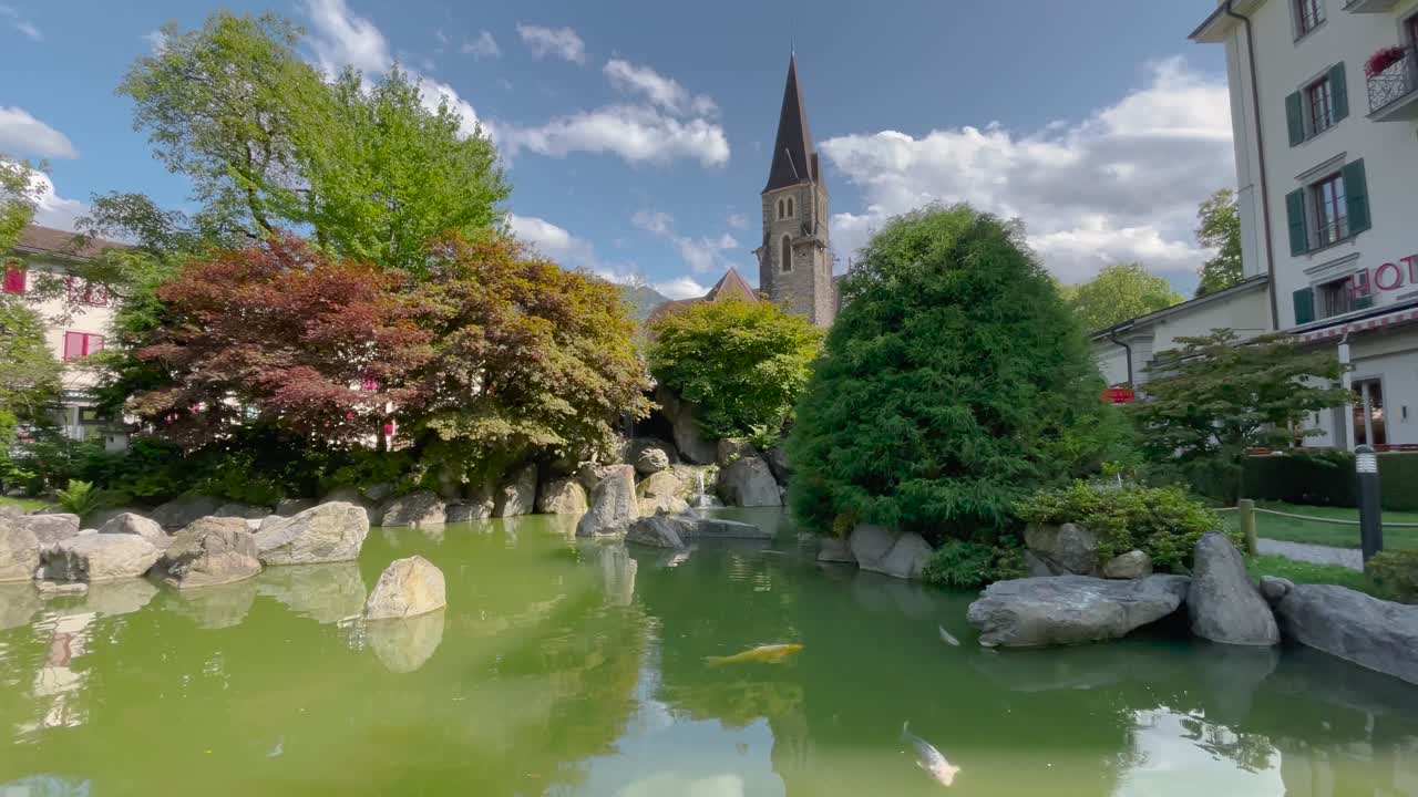 晴れて一部曇りの日の静止画。緑豊かな日本庭園の池で色鮮やかな錦鯉が泳ぎ、豊かな木々の後ろには教会の尖塔がそびえる。スイス、インターラーケン。穏やかな雰囲気。