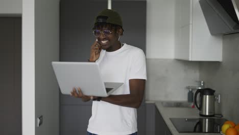Hombre-Afroamericano-Trabajando-En-Casa-En-La-Cocina-Hablando-Por-Teléfono-Inteligente-Y-Usando-Una-Computadora-Portátil