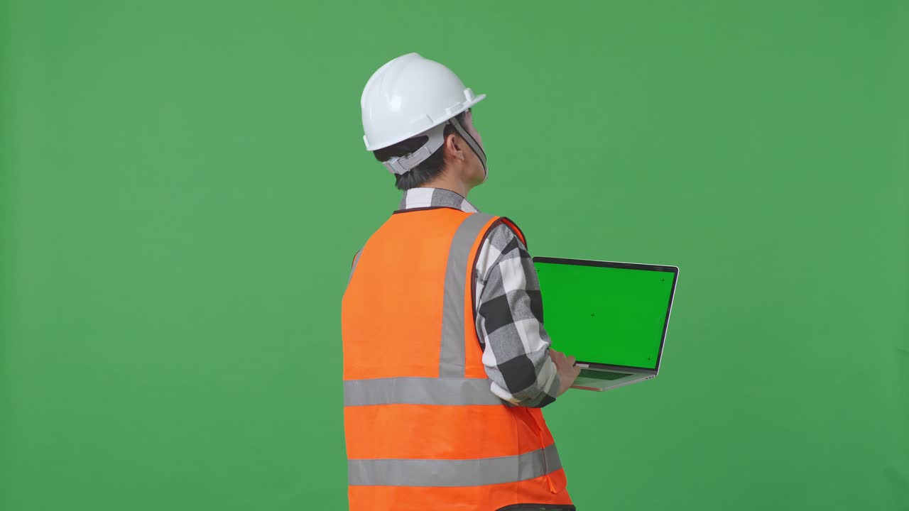 Back View Of Asian Male Engineer With Safety Helmet Working On A Green Screen Laptop And Looking Around While Standing In The Green Screen Background Studio