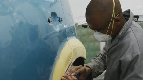 African-American-male-car-mechanic-wearing-a-face-mask-and-using-a-polisher-on-the-side-of-a-car