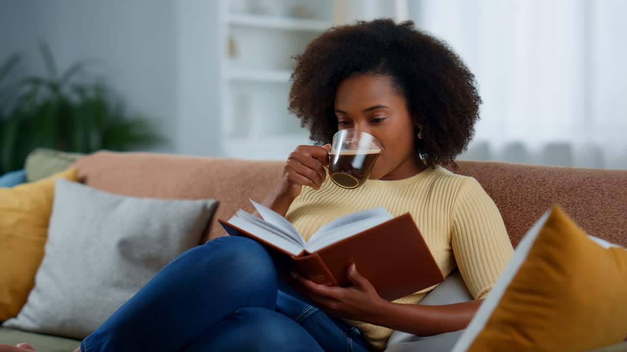 Junge schwarze Frau liest ein Buch und trinkt Kaffee auf einem Sofa