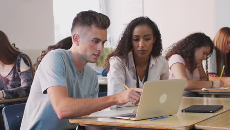 tutora femenina dando apoyo uno a uno al estudiante masculino que trabaja en el escritorio en la computadora portátil